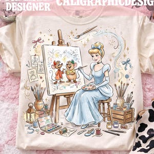 Puede incluir: Camiseta color crema con una ilustración de Cenicienta. El diseño muestra a Cenicienta pintando dos ratones. El texto "ORIGINAL DESIGNER CALIGRAPHICDESIGN" está impreso en la parte superior. La camiseta se muestra sobre una superficie rosa.