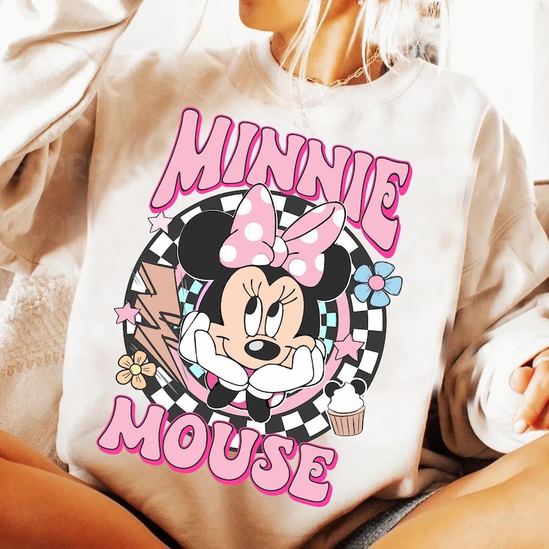 May include: Cream-colored sweatshirt featuring a graphic of Minnie Mouse. The design includes Minnie's face, a pink bow with white polka dots, and the words "MINNIE MOUSE" in pink bubble letters. Additional elements include flowers, stars, and a cupcake.