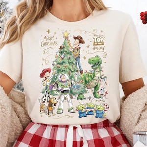 May include: Cream-colored t-shirt featuring a festive "Toy Story Holiday" design. The graphic includes Woody placing a star on a Christmas tree, with Buzz Lightyear, Rex, and other characters. The words "Merry Christmas" are also displayed.