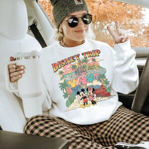 May include: White sweatshirt with a colorful "DISNEY TRIP" graphic featuring Mickey and Minnie Mouse, a castle, and a train. The person is wearing a gray beanie, sunglasses, and plaid pants. A white tumbler is held in the left hand.