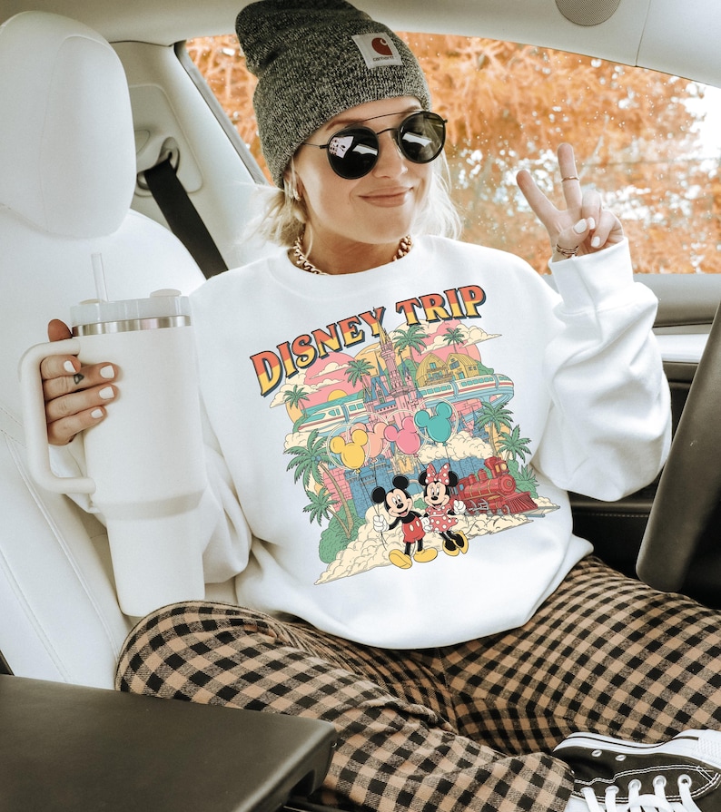 May include: White sweatshirt with a colorful "DISNEY TRIP" graphic featuring Mickey and Minnie Mouse, a castle, and a train. The person is wearing a gray beanie, sunglasses, and plaid pants. A white tumbler is held in the left hand.