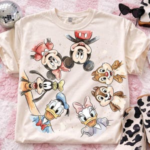 May include: Cream-colored t-shirt featuring a colorful illustration of Disney characters, including Mickey Mouse, Minnie Mouse, Donald Duck, Daisy Duck, Goofy, and Chip and Dale. The design is surrounded by small star accents.