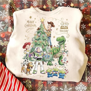 May include: Off-white sweatshirt featuring a colorful graphic of Toy Story characters decorating a Christmas tree. The design includes Woody, Buzz Lightyear, Rex, and other characters, with the text "Merry Christmas" and "Toy Story Holiday".