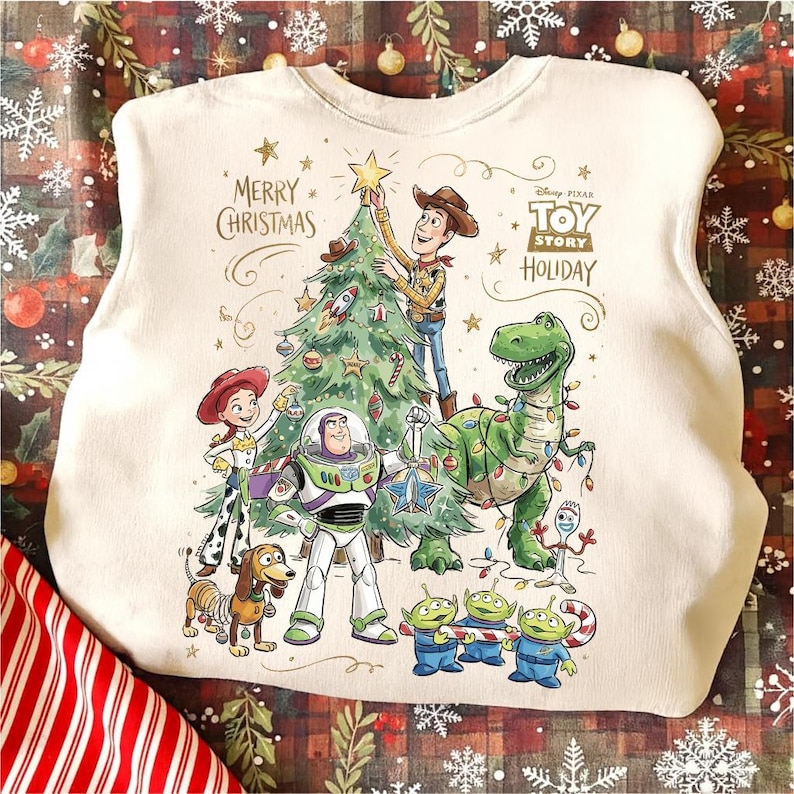 May include: Off-white sweatshirt featuring a colorful graphic of Toy Story characters decorating a Christmas tree. The design includes Woody, Buzz Lightyear, Rex, and other characters, with the text "Merry Christmas" and "Toy Story Holiday".