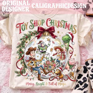 May include: A cream-colored t-shirt featuring a graphic of a Christmas ornament containing Toy Story characters including Woody, Buzz Lightyear, Jessie, Rex, and Slinky Dog. The text reads Toy Shop Christmas and Merry, Bright & Full of Magic.