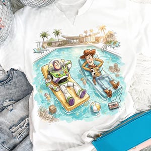 May include: White t-shirt featuring a colorful illustration of Buzz Lightyear and Woody relaxing by a pool. The design includes a pool, palm trees, and a mid-century modern house. The text "VARELOSTUDIO" is at the top.