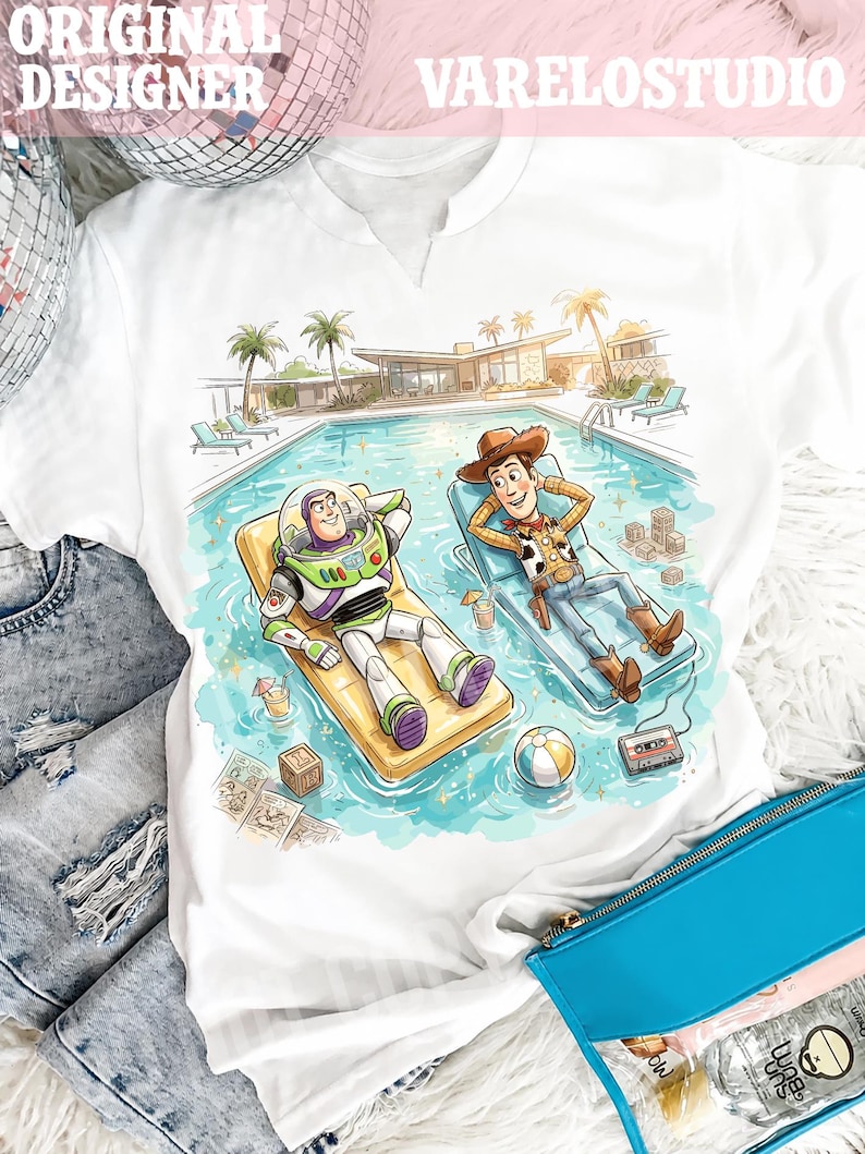 May include: White t-shirt featuring a colorful illustration of Buzz Lightyear and Woody relaxing by a pool. The design includes a pool, palm trees, and a mid-century modern house. The text "VARELOSTUDIO" is at the top.