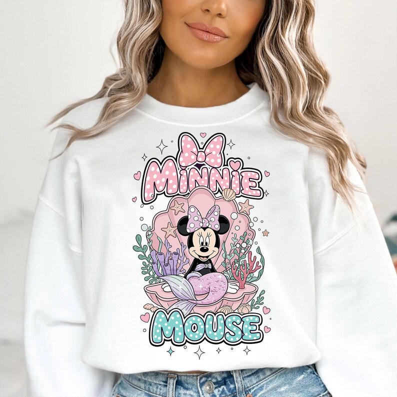 May include: White sweatshirt featuring a Minnie Mouse mermaid design. The graphic includes Minnie in a seashell, with the words "Minnie Mouse" in pink and blue bubble letters. The design has a cartoon style.