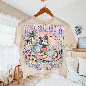 May include: Cream-colored t-shirt featuring a graphic of Mickey Mouse surfing. The design includes the text "Beach Bum", palm trees, waves, butterflies, and flowers. The shirt is hanging on a wooden hanger.