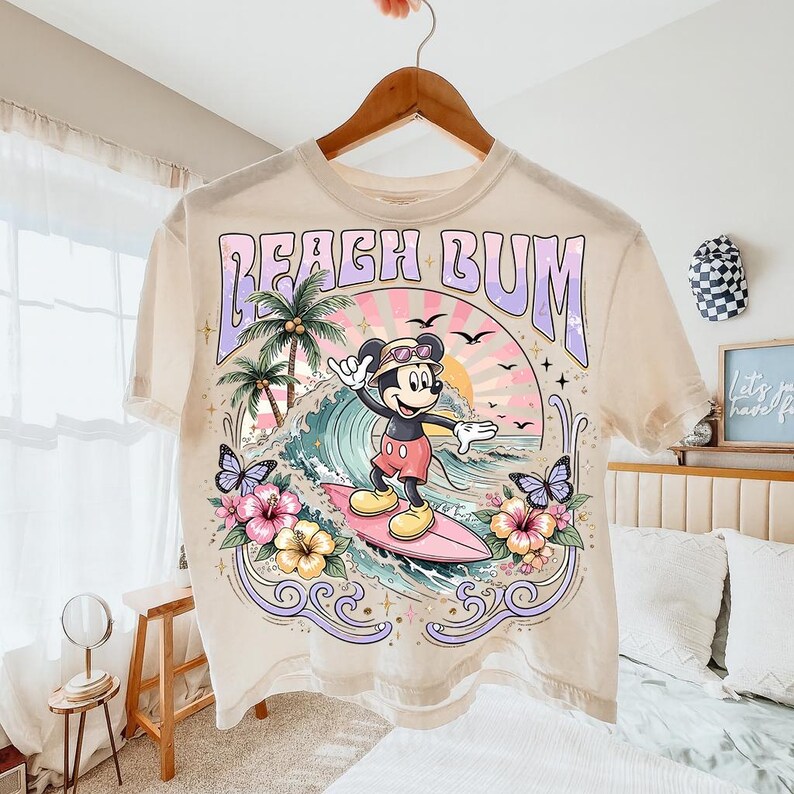 May include: Cream-colored t-shirt featuring a graphic of Mickey Mouse surfing. The design includes the text "Beach Bum", palm trees, waves, butterflies, and flowers. The shirt is hanging on a wooden hanger.