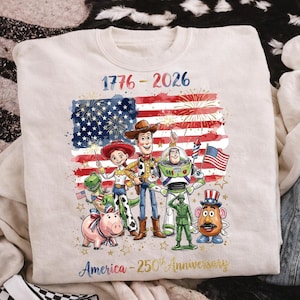 K&ouml;nnte beinhalten: Cremefarbenes Sweatshirt mit einer farbenfrohen Grafik der amerikanischen Flagge, Feuerwerk und Zeichentrickfiguren. Der Text "1776-2026" und "America - 250th Anniversary" sind ebenfalls abgebildet. Das Design zeigt Figuren aus Toy Story.