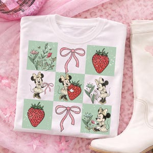May include: White t-shirt featuring a grid design with Minnie Mouse illustrations, strawberries, and pink bows. The shirt has a crew neck and is paired with a white boot on a pink background. The design includes floral elements.