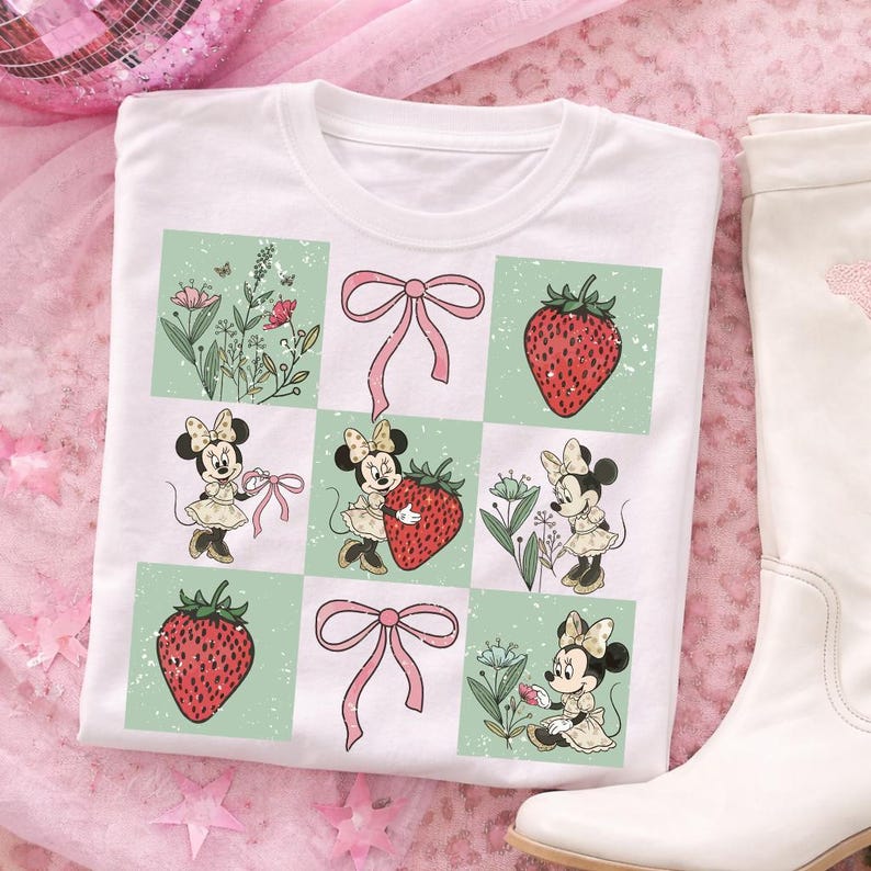 May include: White t-shirt featuring a grid design with Minnie Mouse illustrations, strawberries, and pink bows. The shirt has a crew neck and is paired with a white boot on a pink background. The design includes floral elements.