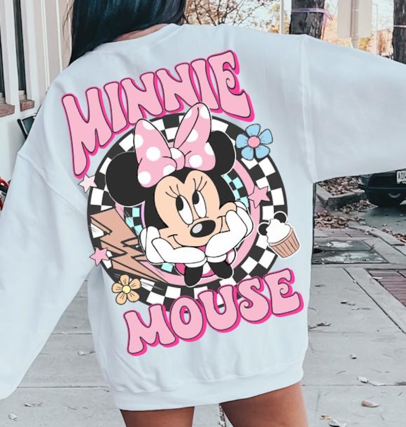 May include: White sweatshirt featuring a colorful graphic of Minnie Mouse. The design includes the words "MINNIE MOUSE" in pink bubble letters, a polka-dot bow, and a cupcake. The graphic is set against a black and white checkered background.