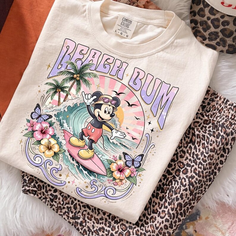 May include: Cream-colored t-shirt with a graphic of Mickey Mouse surfing, the words "Beach Bum", palm trees, and butterflies. The shirt is paired with leopard print pants. The shirt has a Comfort Colors tag.