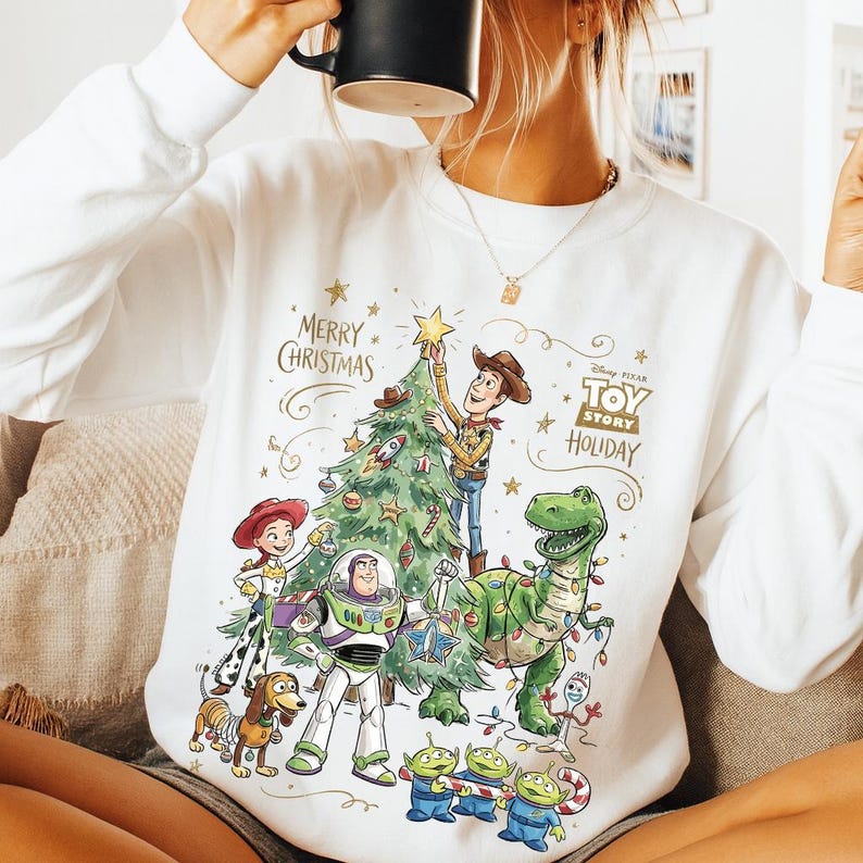 May include: White sweatshirt featuring a colorful graphic of Toy Story characters decorating a Christmas tree. The design includes Woody, Buzz Lightyear, Rex, and the Little Green Men, with the text "MERRY CHRISTMAS" and "TOY STORY HOLIDAY".
