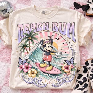 May include: Cream-colored t-shirt with a retro graphic featuring Mickey Mouse surfing. The design includes the words "Beach Bum," palm trees, waves, butterflies, and flowers. The shirt has a crew neck and short sleeves.