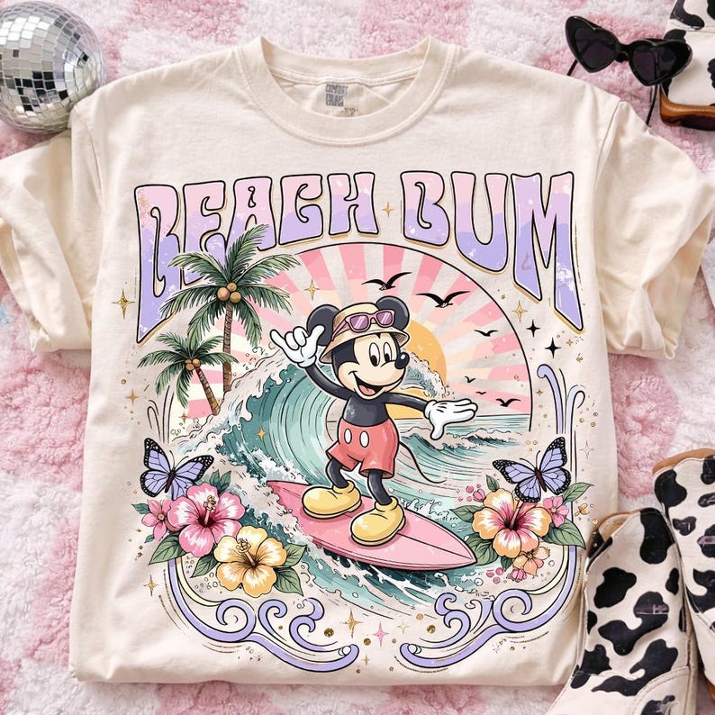 May include: Cream-colored t-shirt with a retro graphic featuring Mickey Mouse surfing. The design includes the words "Beach Bum," palm trees, waves, butterflies, and flowers. The shirt has a crew neck and short sleeves.