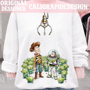 Può includere: Felpa bianca con una grafica di Woody, Buzz Lightyear e i piccoli omini verdi di Toy Story. Il design include un artiglio meccanico e il testo "ORIGINAL DESIGNER CALIGRAPHICDESIGN".