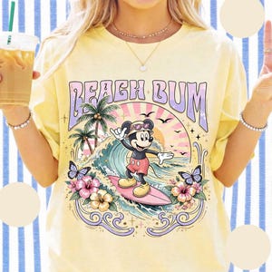May include: Yellow t-shirt with a graphic of Mickey Mouse surfing, the text "BEACH BUM", palm trees, waves, butterflies, and flowers. The design features a retro, vintage style with a pastel color palette.