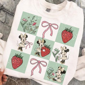 May include: White sweatshirt featuring a grid design with Minnie Mouse illustrations, strawberries, pink bows, and floral elements. The design has a vintage, distressed look. The sweatshirt is a casual, comfortable garment.