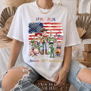 K&ouml;nnte beinhalten: Wei&szlig;es T-Shirt mit einer Grafik der amerikanischen Flagge, Feuerwerk und Toy Story-Charakteren. Das Shirt zeigt den Text "1776-2026" und "America - 250th Anniversary". Ein patriotisches Design.