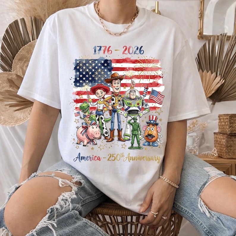 K&ouml;nnte beinhalten: Wei&szlig;es T-Shirt mit einer Grafik der amerikanischen Flagge, Feuerwerk und Toy Story-Charakteren. Das Shirt zeigt den Text "1776-2026" und "America - 250th Anniversary". Ein patriotisches Design.