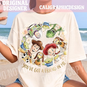 以下が含まれることがあります： クリーム色のTシャツ。映画「トイ・ストーリー」のキャラクター、ウッディ、バズ・ライトイヤー、ジェシーなどのカラフルなグラフィックデザイン。デザインには、金色の文字で「You've Got a Friend in Me」と書かれています。「ORIGINAL DESIGNER」と「CALIGRAPHICDESIGN」の文字も表示されています。