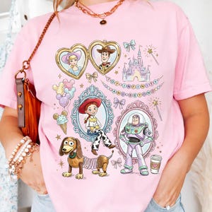 Może przedstawiać: Jasnor&oacute;żowa koszulka z ilustracjami postaci z Toy Story. Projekt zawiera portrety Woody'ego, Buzza Astrala, Jessie i Bo Peep, zamek, balony i frazę "TO INFINITY AND BEYOND."