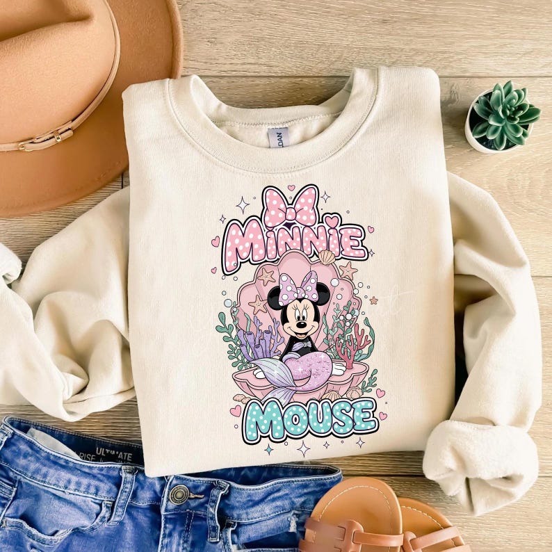 May include: Cream-colored sweatshirt featuring a Minnie Mouse mermaid design. The graphic includes the words "Minnie Mouse" in pink and blue, with Minnie in a seashell. The design is surrounded by coral and stars.