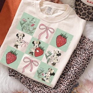 May include: Cream-colored t-shirt featuring a grid pattern with Minnie Mouse illustrations, strawberries, and pink bows. The shirt is paired with leopard print pants and a matching hat. The tag reads "Comfort Colors".