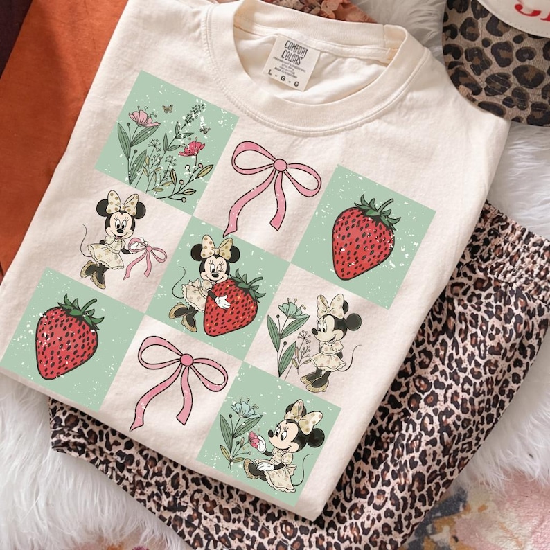 May include: Cream-colored t-shirt featuring a grid pattern with Minnie Mouse illustrations, strawberries, and pink bows. The shirt is paired with leopard print pants and a matching hat. The tag reads "Comfort Colors".