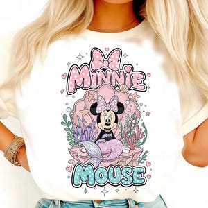 May include: White t-shirt featuring a Minnie Mouse graphic. Minnie is depicted as a mermaid inside a seashell, with the words "Minnie Mouse" above and below. The design includes pink, purple, and teal colors.