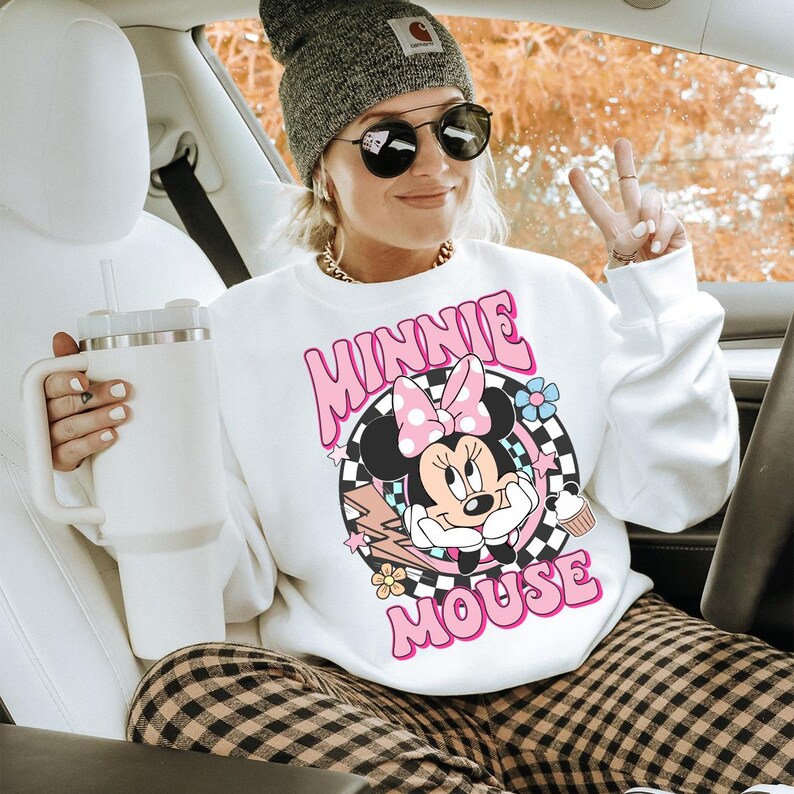 May include: White sweatshirt featuring a Minnie Mouse graphic with a pink bow, set against a checkered background. The sweatshirt has the words "MINNIE MOUSE" in pink. The person is wearing a gray beanie, sunglasses, and holding a white tumbler.