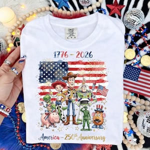K&ouml;nnte beinhalten: Wei&szlig;es T-Shirt mit patriotischem Design. Der Text lautet "1776-2026" und "America - 250th Anniversary." Die Grafik enth&auml;lt eine amerikanische Flagge und Zeichentrickfiguren. Das Shirt ist von der Marke Comfort Colors.