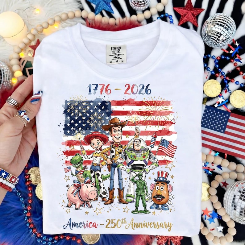 K&ouml;nnte beinhalten: Wei&szlig;es T-Shirt mit patriotischem Design. Der Text lautet "1776-2026" und "America - 250th Anniversary." Die Grafik enth&auml;lt eine amerikanische Flagge und Zeichentrickfiguren. Das Shirt ist von der Marke Comfort Colors.