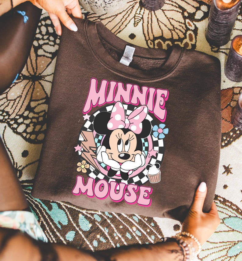 May include: A brown sweatshirt featuring a graphic of Minnie Mouse with a pink bow and the text "MINNIE MOUSE" in pink. The design includes a checkered background, flowers, and a cupcake.