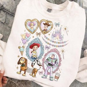Może przedstawiać: Biała bluza z kolorowymi ilustracjami postaci z Toy Story, w tym Chudego, Buzz'a Astrala i Jessie, w ozdobnych ramkach. Projekt zawiera napis "TO INFINITY AND BEYOND" oraz inne elementy dekoracyjne.