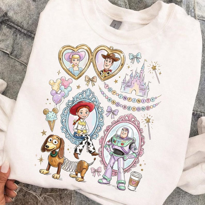 Może przedstawiać: Biała bluza z kolorowymi ilustracjami postaci z Toy Story, w tym Chudego, Buzz'a Astrala i Jessie, w ozdobnych ramkach. Projekt zawiera napis "TO INFINITY AND BEYOND" oraz inne elementy dekoracyjne.