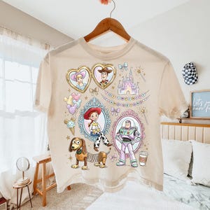 Może przedstawiać: Kremowa koszulka z kolorową grafiką postaci z filmu Toy Story. Projekt zawiera portrety w ozdobnych ramach, zamek i napis "TOY STORY AND BEYOND". Koszulka wisi na drewnianym wieszaku.