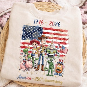 K&ouml;nnte beinhalten: Cremefarbenes Sweatshirt mit patriotischem Design. Es zeigt die amerikanische Flagge, Feuerwerk und Zeichentrickfiguren. Der Text lautet "1776-2026" und "America - 250th Anniversary".