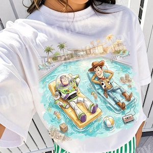 May include: White t-shirt featuring a colorful illustration of Buzz Lightyear and Woody relaxing by a pool. The design includes palm trees, a modern house, and the text "ORIGINAL DESIGNER CALIGRAPHICDESIGN".