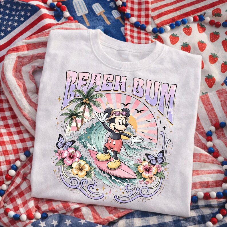 May include: White t-shirt featuring a colorful graphic of Mickey Mouse surfing. The design includes the text "BEACH BUM", palm trees, waves, butterflies, and flowers. The shirt is surrounded by patriotic decorations.