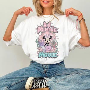 May include: White t-shirt featuring a Minnie Mouse graphic. The design includes Minnie as a mermaid, surrounded by aquatic elements, with the words "Minnie Mouse" in a playful font. The shirt has a classic crew neck and short sleeves.