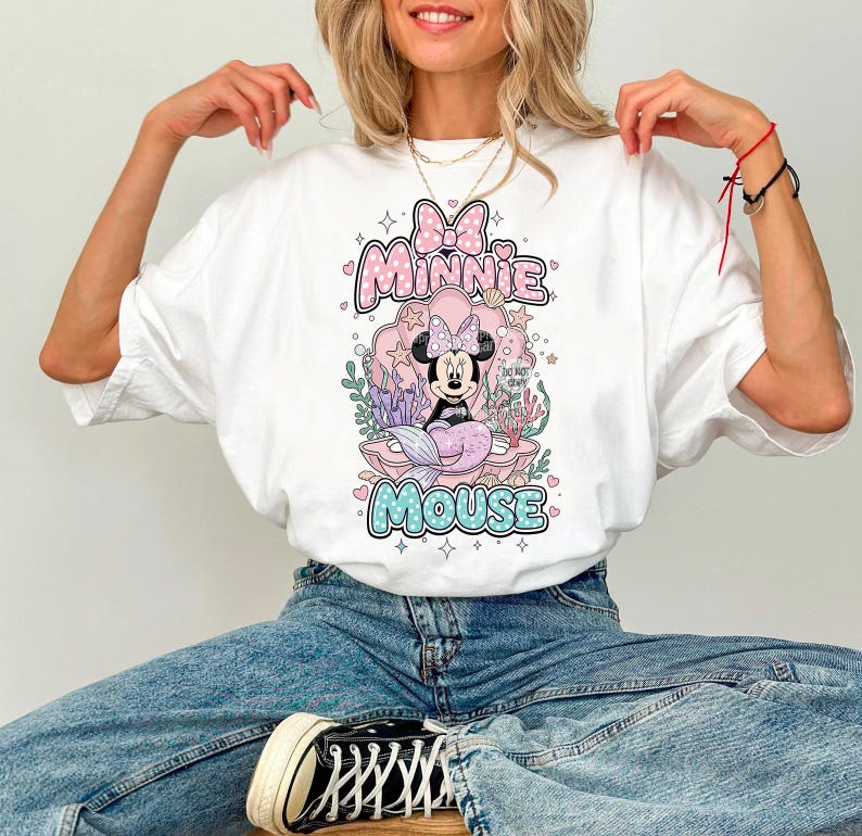 May include: White t-shirt featuring a Minnie Mouse graphic. The design includes Minnie as a mermaid, surrounded by aquatic elements, with the words "Minnie Mouse" in a playful font. The shirt has a classic crew neck and short sleeves.