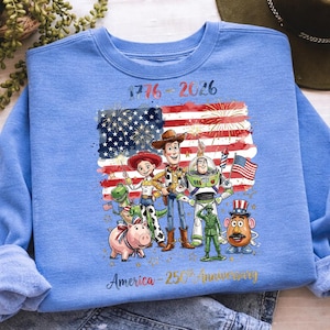 K&ouml;nnte beinhalten: Hellblaues Sweatshirt mit einer Grafik von Zeichentrickfiguren vor einer amerikanischen Flagge mit Feuerwerk. Der Text lautet "1776-2026" und "America - 250th Anniversary" in Gold.