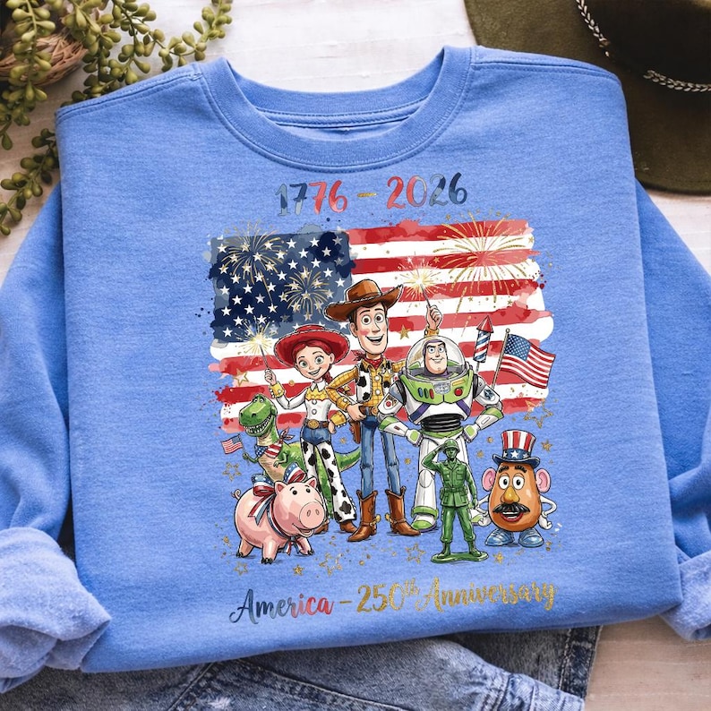 K&ouml;nnte beinhalten: Hellblaues Sweatshirt mit einer Grafik von Zeichentrickfiguren vor einer amerikanischen Flagge mit Feuerwerk. Der Text lautet "1776-2026" und "America - 250th Anniversary" in Gold.