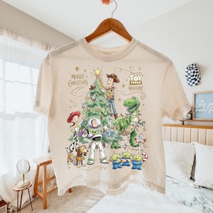 May include: Cream-colored t-shirt featuring a festive Toy Story holiday design. The graphic includes Woody, Buzz Lightyear, Rex, and other characters decorating a Christmas tree. Text reads "Merry Christmas" and "Toy Story Holiday".