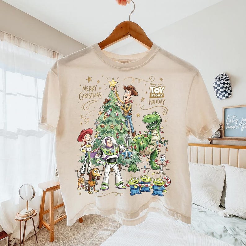 May include: Cream-colored t-shirt featuring a festive Toy Story holiday design. The graphic includes Woody, Buzz Lightyear, Rex, and other characters decorating a Christmas tree. Text reads "Merry Christmas" and "Toy Story Holiday".
