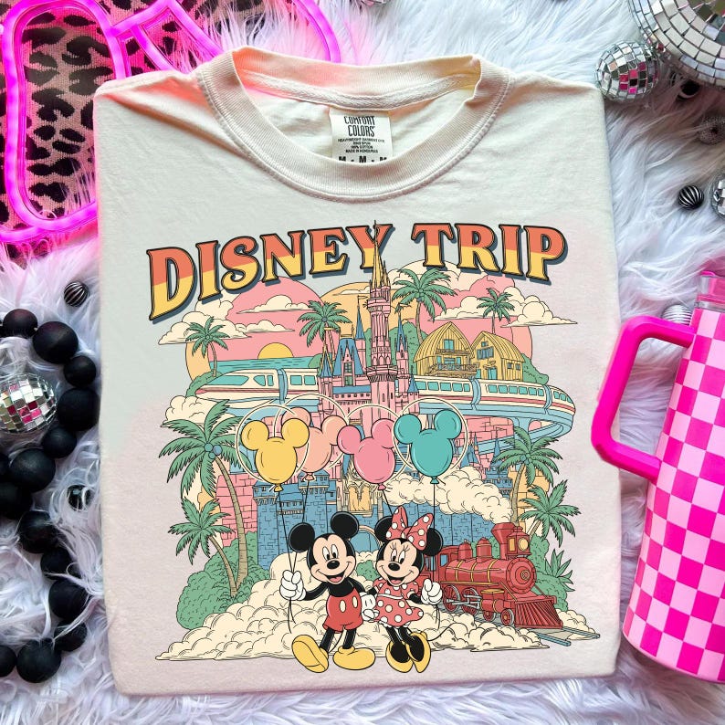 May include: Cream-colored t-shirt with a colorful "DISNEY TRIP" graphic. The design features Mickey and Minnie Mouse, a castle, palm trees, balloons, and a train. The shirt is a Comfort Colors brand.